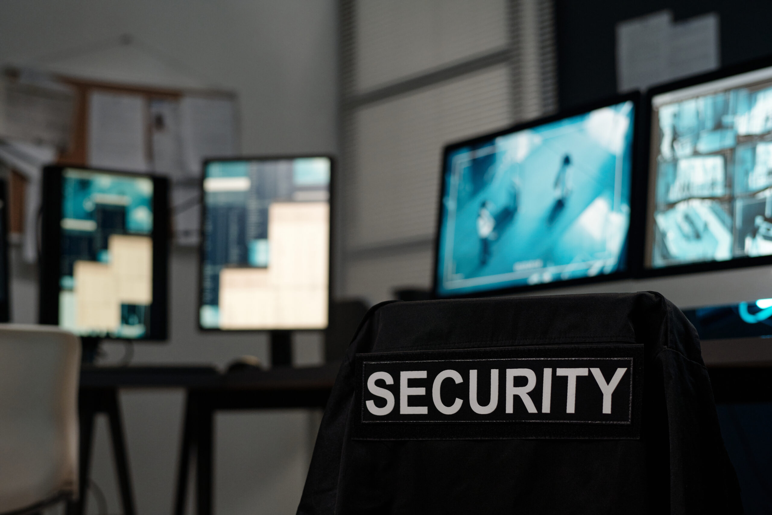 Black uniform with word security hanging on empty chair standing by workplace of officer with several computer monitors