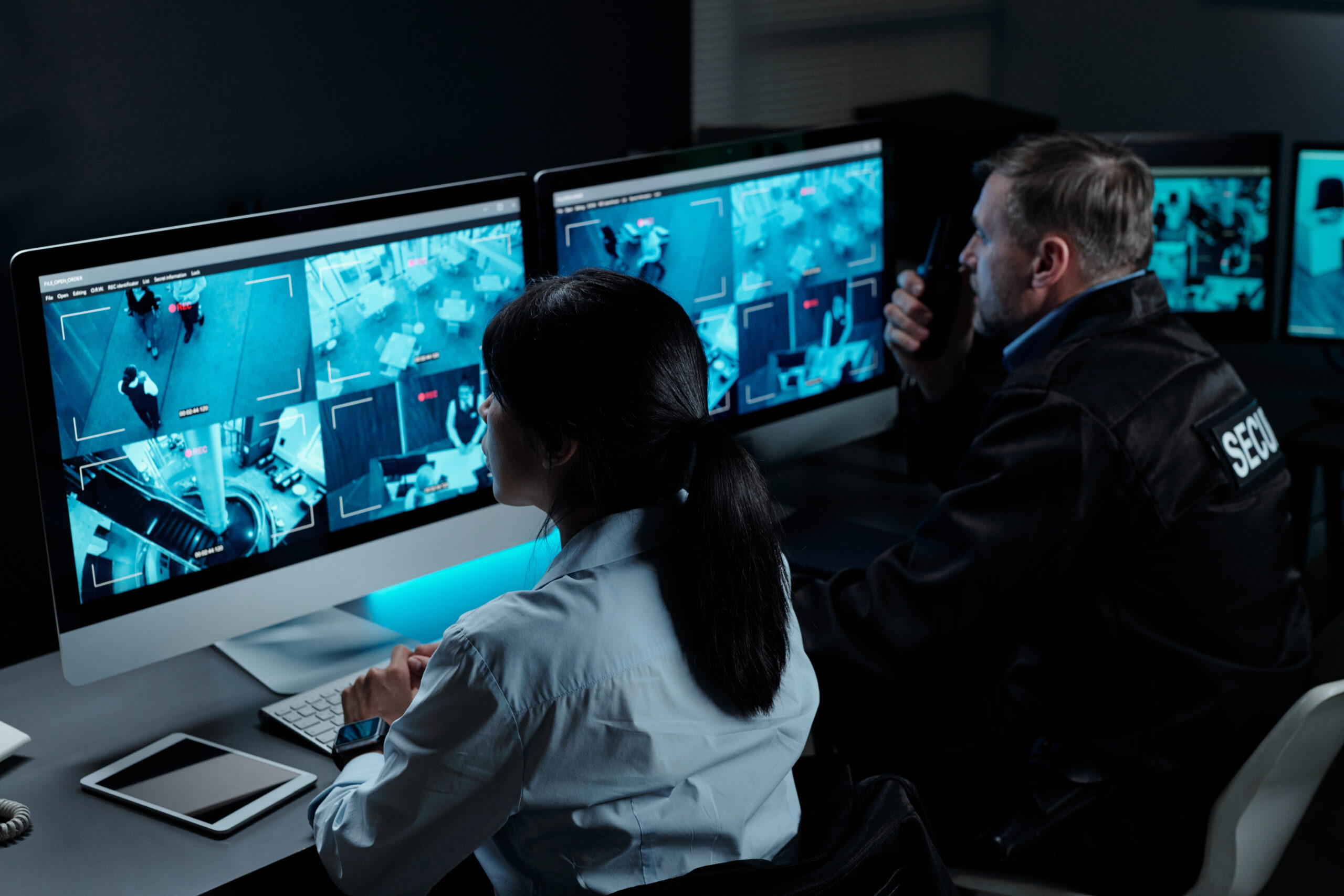Two intercultural male and female officers in uniform sitting in front of computer monitors with cctv video system on screens