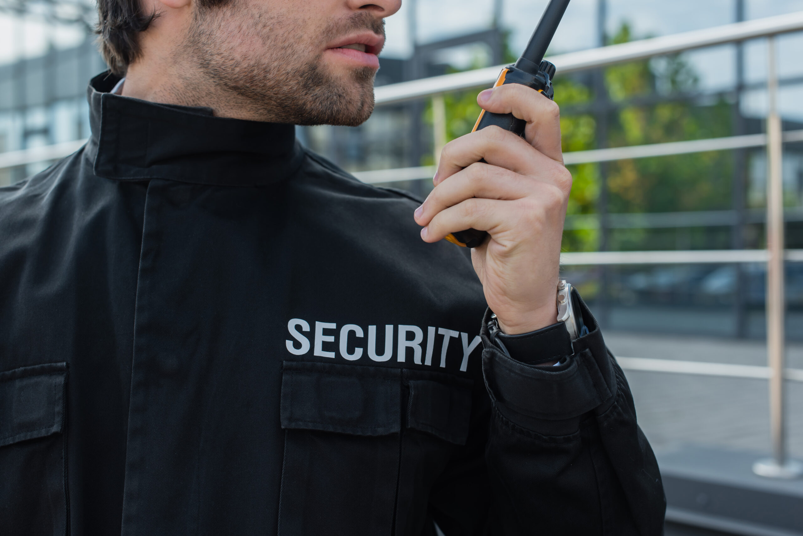 partial view of guard in uniform with security lettering talking on walkie-talkie outdoors